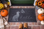 A dark charcoal doormat with a gray cat, butterfly, and 'hello' script, placed on a brick porch in front of a green door. Pumpkins, sunflowers, a lantern, and a person's tan boots are visible around it.