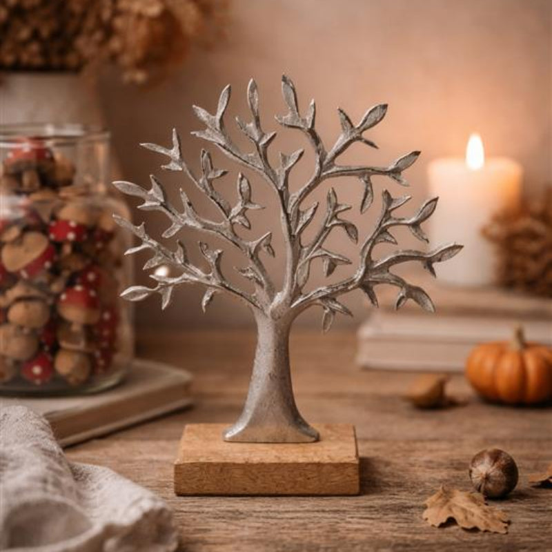 A decorative silver-toned metal tree sculpture with a textured trunk and leafless branches, mounted on a rectangular wooden base, displayed on a rustic wooden table with candles, dried leaves, and small pumpkins in the background.