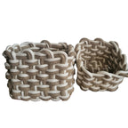 Two woven storage baskets with a braided jute and white rope pattern, displayed on a wooden chest against a white wall.