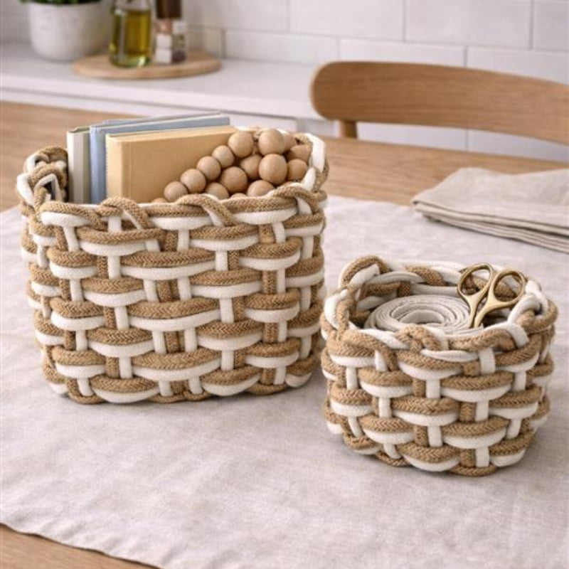 Two woven rope storage baskets in a kitchen setting, one larger basket holding books and wooden beads and one smaller basket holding a tin, placed on a light linen runner over a wooden table.