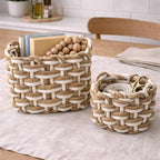 Two woven rope storage baskets in a kitchen setting, one larger basket holding books and wooden beads and one smaller basket holding a tin, placed on a light linen runner over a wooden table.