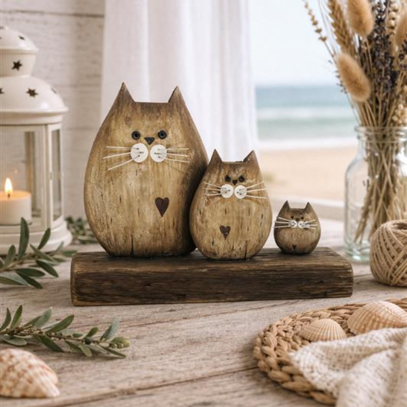 Three rustic wooden cat figurines in different sizes, arranged on a dark wooden block on a weathered wooden table. The scene is styled with a lantern, dried grass in a glass jar, and a knitted blanket, with a soft-focus beach background visible through a window.