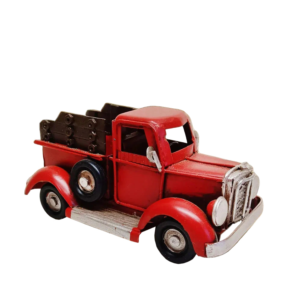 A small red vintage toy pickup truck with black wheels and silver grille details, shown on a wooden surface with a blurred indoor background featuring plants and a yellow wall.